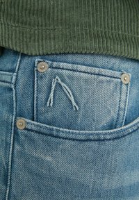 Vaaleansiniset farkut, joissa takataskussa näkyvä ommeltu logo ja metalliset napit, yhdistetty teksturoituun vihreään corduroypaitaan.