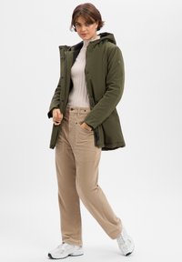 Parka vert olive avec capuche, fermeture par boutons et zip, portée sur un pull léger côtelé et un pantalon en velours côtelé beige. Baskets blanches avec des accents.
