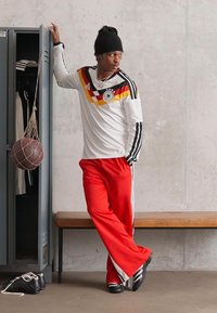 Λευκή μακριά μπλούζα με κόκκινες, κίτρινες και μαύρες ρίγες; κόκκινες παντελόνες; μαύρο σκουφάκι; μαύρα παπούτσια; ντουλάπα και μπάλα μπάσκετ στο φόντο.