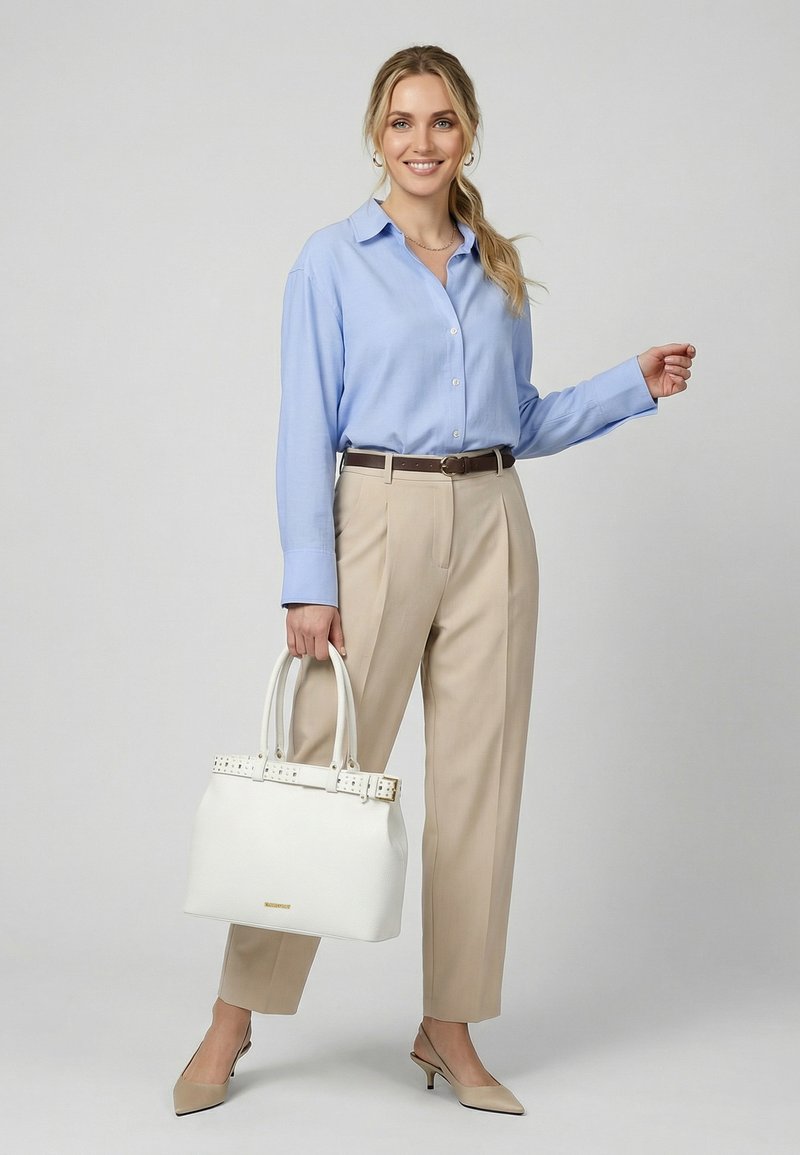 Femme souriante, portant une chemise bleu clair, un pantalon beige et des talons beiges, tenant un sac à main blanc sur un fond clair uni.