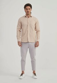 Homme portant une chemise beige à rayures à manches longues, un pantalon court gris clair et des chaussures blanches à enfiler, se tenant contre un fond blanc.
