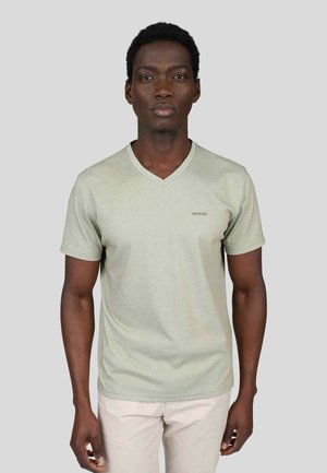 T-shirt vert clair à manches courtes en V en tissu doux avec un détail de logo subtil sur la poitrine. Le modèle pose de face contre un fond gris.