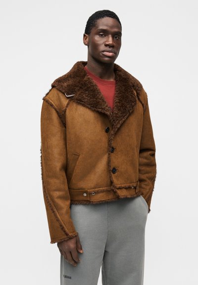 Καφέ μπουφάν από shearling με μεγάλο γιακά, κλείσιμο με κουμπιά και φθαρμένες άκρες· φορεμένο πάνω από κόκκινο πουκάμισο και γκρι φόρμες.