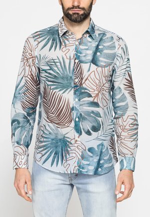 Uomo che indossa una camicia a maniche lunghe con stampa a foglie tropicali blu e marroni e jeans chiari slavati.