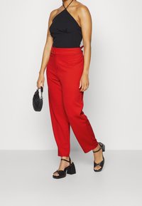 Rode high-waisted broek met een tapse pasvorm, gecombineerd met een zwarte haltertop. Accessoires zijn een kleine zwarte handtas en chunky zwarte hakschoenen.