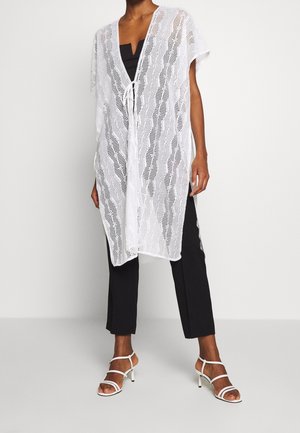 Mujer con un poncho blanco de punto abierto con lazo delantero, pantalones negros y sandalias de tacón blancas con tiras, de pie frente a un fondo liso.