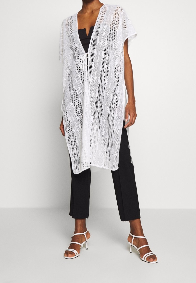 Femme portant un poncho blanc en maille ouverte avec un lien à l'avant, un pantalon noir et des sandales blanches à talons avec des brides, debout devant un fond uni.