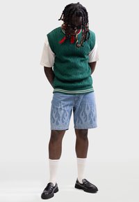 Uomo con dreadlocks che indossa un gilet verde lavorato a maglia con cavi, una maglietta bianca, pantaloncini di jeans con motivo a fiamme, sciarpa rossa al collo, calzini bianchi e mocassini neri, in piedi.