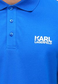 Modra polo majica s klasičnim ovratnikom, tremi modrimi gumbi in belim logotipom z napisom "KARL LAGERFELD" na levi strani. Gladek tekstura.