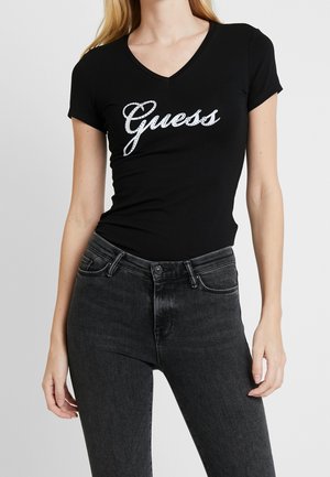 Kvinde iført en tætsiddende sort V-hals T-shirt med "Guess" funklende logo og højttaljede mørkegrå jeans, stående mod en ensfarvet baggrund.