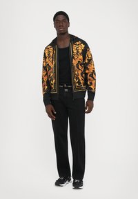 Jeune homme debout, portant une veste zippée à motif noir et or, un débardeur noir, un pantalon noir et des baskets noir et blanc, sur fond blanc.