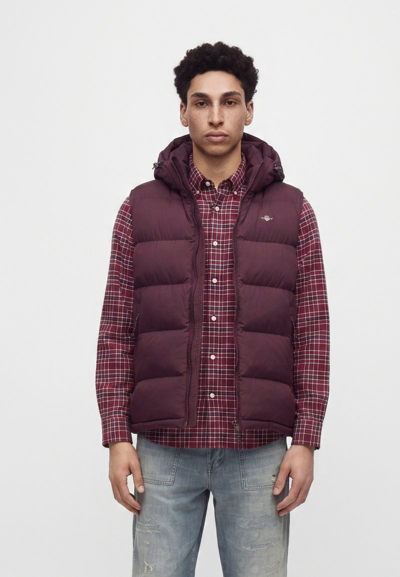 Jeune homme portant une doudoune bordeaux par-dessus une chemise à carreaux rouge et un jean bleu clair déchiré, debout devant un fond uni.