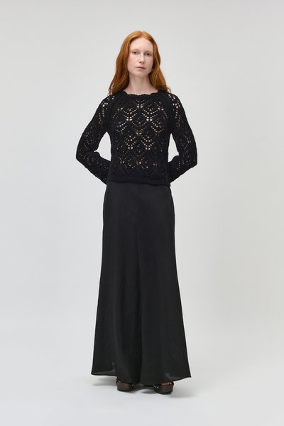 Femme rousse debout, les mains derrière le dos, portant un top noir en crochet à manches longues et une longue jupe noire avec des chaussures à plateforme.