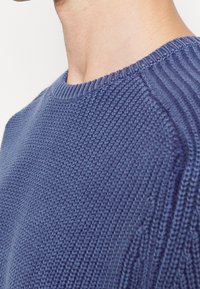 Gros plan d'une personne portant un pull en tricot bleu montrant une texture détaillée et un col ainsi que des épaules côtelés.