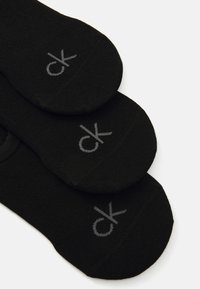 Schwarze Knöchelsocken mit einer glatten Textur, die ein graues "ck"-Logo auf der Oberseite tragen. Drei Paare übereinander gestapelt.