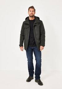 Man wearing black hooded jacket, black shirt, blue jeans, and black boots standing against white background.