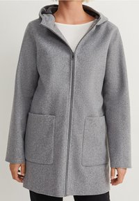 Manteau gris à capuche avec fermeture éclair, deux poches avant, porté par-dessus une chemise blanche et un pantalon noir, visible du cou jusqu'à la mi-cuisse.