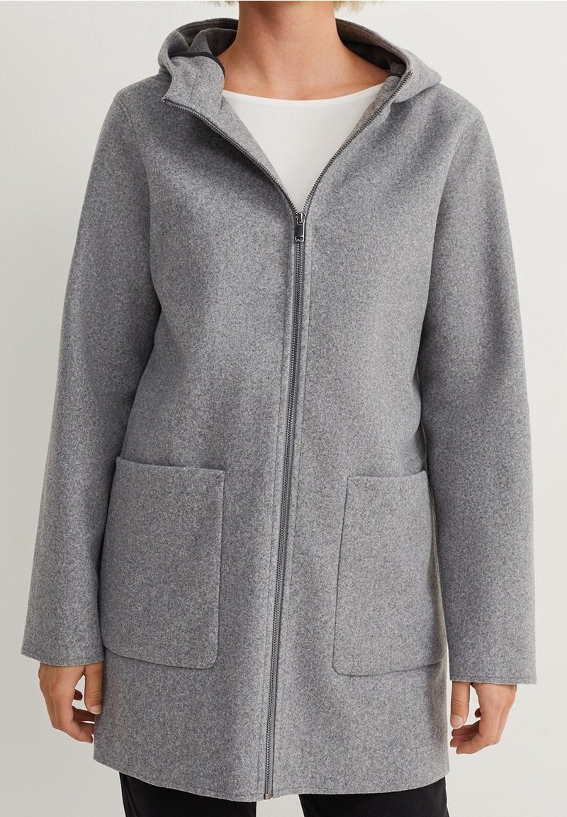 Manteau gris à capuche avec fermeture éclair, deux poches avant, porté par-dessus une chemise blanche et un pantalon noir, visible du cou jusqu'à la mi-cuisse.