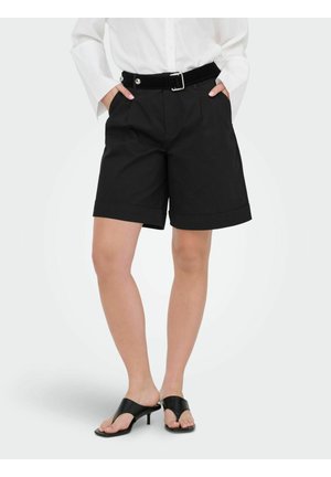 Osoba nosiaca čierne šortky s opaskom po kolená, bielu blúzku s dlhými rukávmi a čierne sandále na opätku, stojaca s rukami v vreckách.