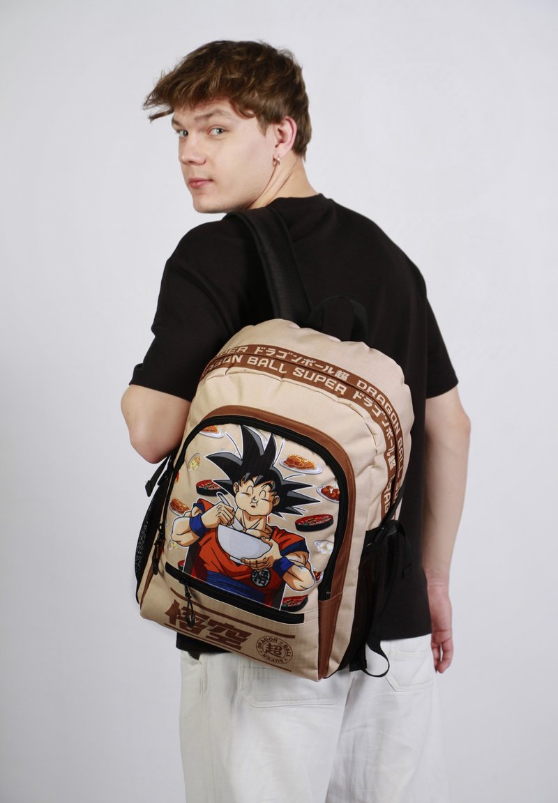Un sac à dos tan avec des bretelles noires comporte un personnage illustré tenant un bol, accentué par des images de nourriture et du texte de marque.