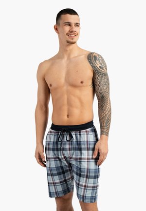 Man zonder shirt met Polynesische tattoo op de rechterarm, die blauwe en witte geruite zwembroek met een zwart koord draagt.