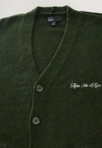 Dunkelgrüner Cardigan aus weichem Strickmaterial mit V-Ausschnitt, zwei Knöpfen und gesticktem "Alpes Côte d'Azur"-Logo in Weiß.