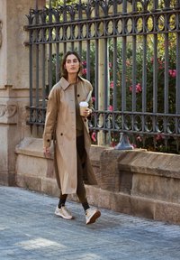 Gabardina beige con botones grandes, llevada sobre un atuendo oscuro. Leggings negros y zapatillas de deporte de color claro. Sostenido una taza de café mientras camina.