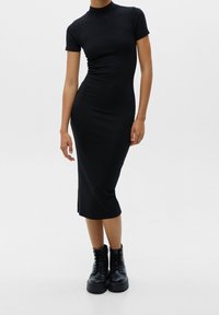 Femme portant une robe midi noire ajustée à manches courtes et col haut, associée à des bottes noires épaisses à lacets.