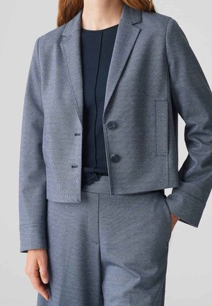 Femme portant un blazer gris court à deux boutons et un pantalon assorti, une main dans la poche, sur un fond uni.