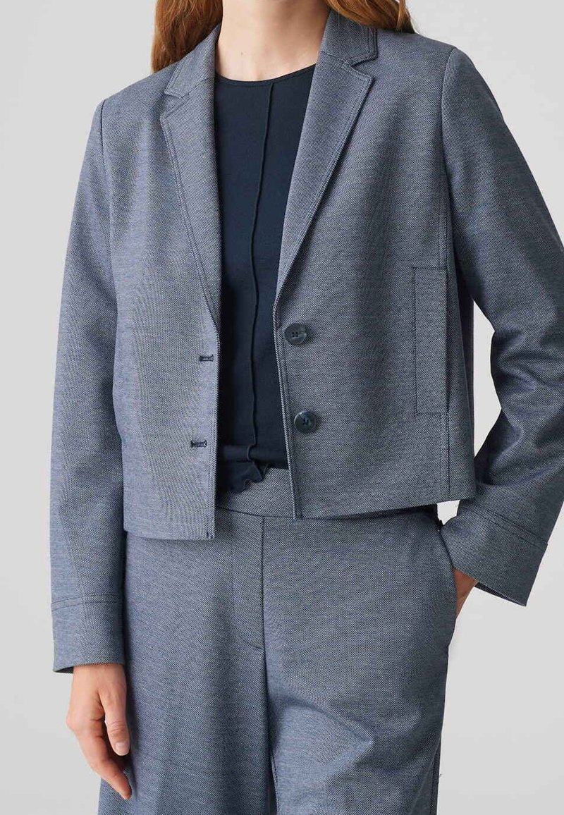 Femme portant un blazer gris court à deux boutons et un pantalon assorti, une main dans la poche, sur un fond uni.