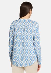 Chemise à manches longues à motifs bleus avec des dessins géométriques en bleu clair, crème et accents dorés. Dispose d'un ourlet arrondi et d'un pli au dos.