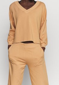 Sudadera corta de color beige claro con escote en V, hombros caídos y puños elásticos, combinada con pantalones de pierna ancha a juego con bolsillos.