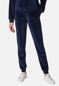 Marineblaue Velours-Jogginghosen mit elastischem Bund, Seitentaschen und schmal zulaufenden Beinen, kombiniert mit weißen Sneakers für einen lässigen Look.