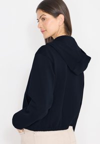 Sweat à capuche court bleu marine avec cordon de serrage, poignets et ceinture côtelés. Texture lisse, design décontracté, idéal pour superposer.