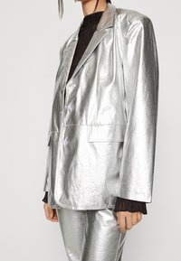Stříbrný metallic blazer s uvolněným střihem, dvěma knoflíky a kapsou. Nosený přes černý texturovaný svetr, který se vyznačuje lesklým vzhledem.