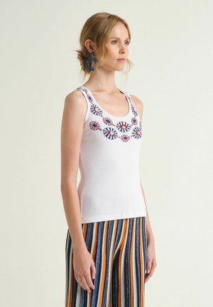 Femme aux cheveux blonds attachés en chignon bas, portant un débardeur blanc orné de motifs d'yeux bleus et rouges, et un pantalon rayé multicolore, debout de profil.