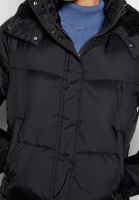 Chaqueta negra acolchada con cierre de cremallera, con cuello de tela texturizada y puños elásticos, dos bolsillos delanteros y detalles acolchados.