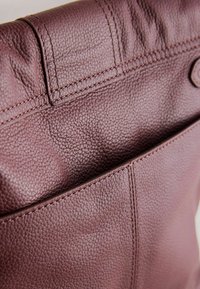 Borsa in pelle bordeaux con superficie texture, caratterizzata da cuciture visibili e chiusura magnetica. Piccolo taschino esterno visibile.
