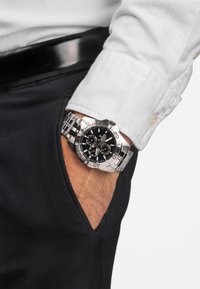 Reloj cronógrafo con una esfera negra, correa de acero inoxidable plateada y marcadores. Lleva puesto en una muñeca, parcialmente visible en un bolsillo.