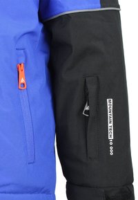 Veste bleu et noire avec une poche zippée dotée d'un tirant orange, des accents réfléchissants et l'inscription "MOUNTAIN TECH 10 000" sur la manche.