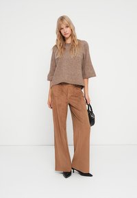 Brauner Strickpullover mit weiten Dreiviertelärmeln, kombiniert mit beigen Wildleder-Hose im weit geschnittenen Stil und schwarzen spitzen High Heels. Kleine schwarze Handtasche.