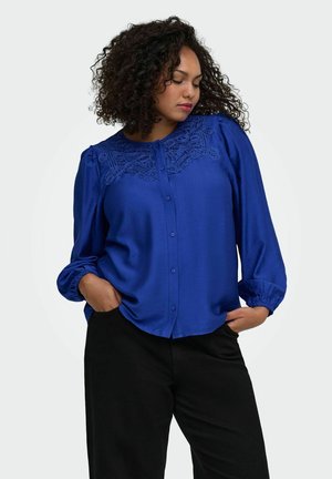 Femme aux cheveux bouclés portant un chemisier bleu roi à manches longues avec un détail en dentelle et un pantalon noir, mains dans les poches, regardant vers le bas sur le côté.