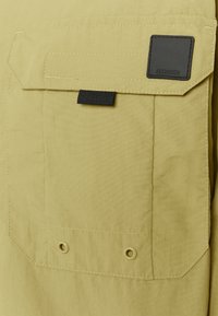 Light yellow fabric with a large patch pocket and a black branding label. Features white stitching and two metal eyelets at the bottom.