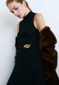 Robe noire sans manches avec un col montant, ornée d'une broche en forme de feuille d'or à la taille. Un châle en fourrure marron est drapé sur une épaule.