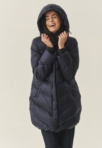 Žena se smiješi s zatvorenim očima, nosi tamnoplavu jaknu s kapuljačom koja joj pokriva glavu, a rukama drži kapuljaču blizu lica.