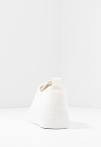 Sneaker bianca in tela con una finitura liscia, punta arrotondata, retro testurizzato e linguetta. Presenta una suola a contrasto e un design minimalista.