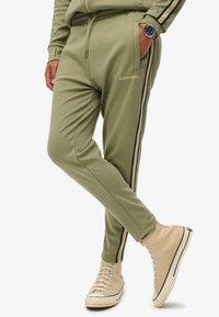 Olijfgroene joggers met zwarte en beige zijstrepen, voorzien van een elastische tailleband en logo-detail. Gekoppeld met beige hoge sneakers.