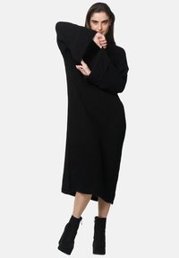 Czarna dzianinowa sukienka swetrowa z szerokimi, dzwonkowatymi rękawami, sięgająca do połowy łydki, luźnego kroju i fakturowanej tkaniny. Zestawiona z czarnymi botkami za kostkę.