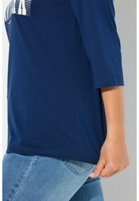 Granatowy długi rękaw z okrągłym dekoltem, miękka tkanina, boczne rozcięcia oraz biały nadruk graficzny z przodu. Noszony z jasnoniebieskimi dżinsami.