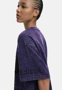 Fialové oversize tričko s obnošenou textúrou, voľným strihom a potlačou značky "DESIGUAL" na rukáve.